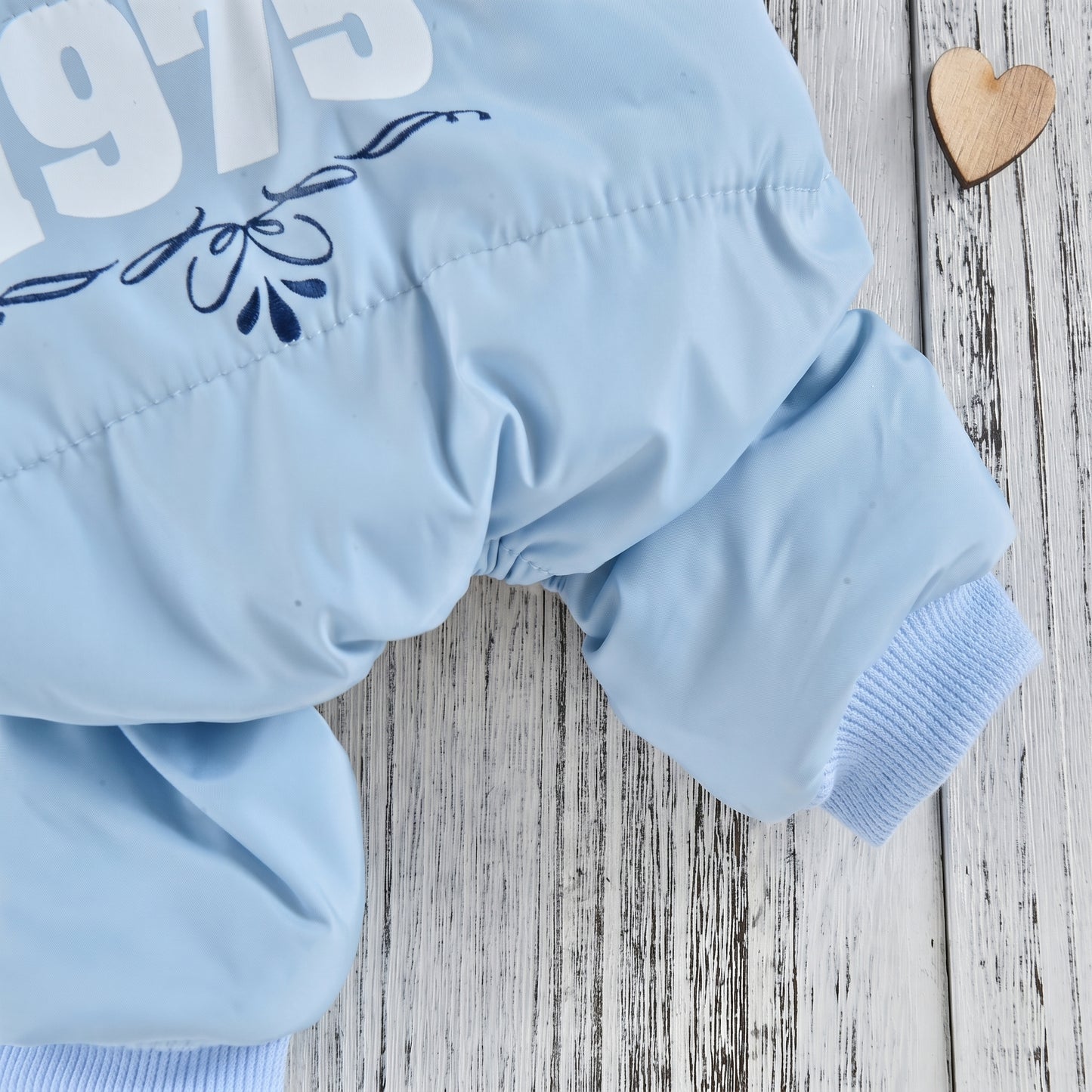 Close-up of a light blue dog jumpsuit leg and cuff, with '1975' print detail, displayed on a white wooden background with a wooden heart ornament.