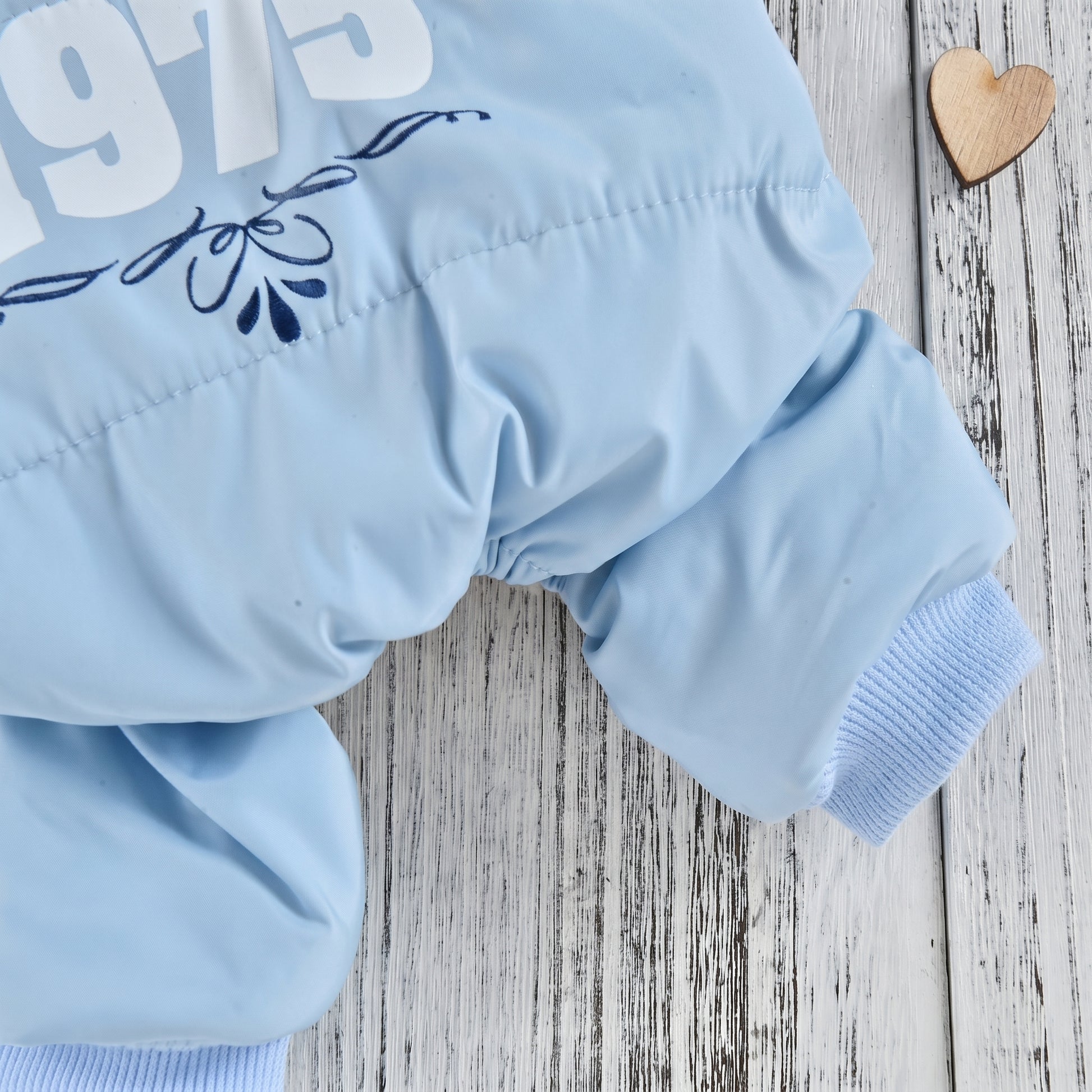 Close-up of a light blue dog jumpsuit leg and cuff, with '1975' print detail, displayed on a white wooden background with a wooden heart ornament.