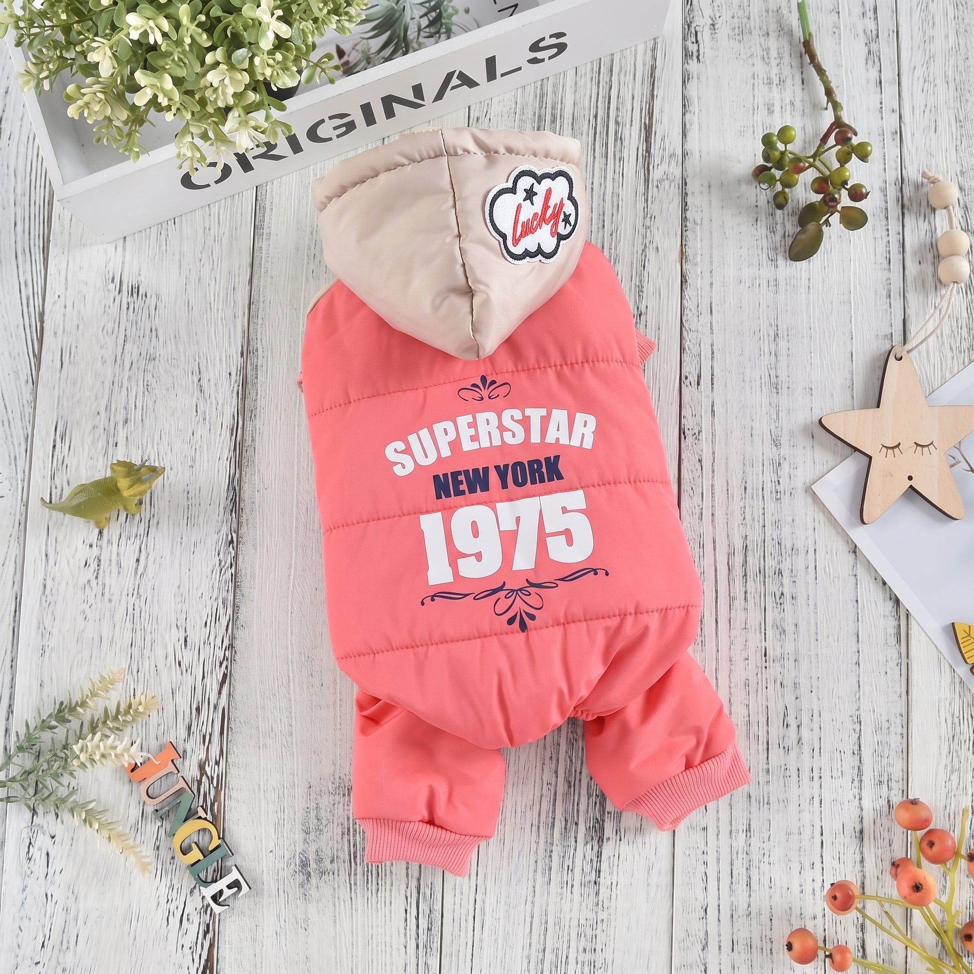 CozyCloud Full-Body pink dog jumpsuit with a hood, featuring 'Superstar New York 1975' print, displayed on a rustic wooden table.