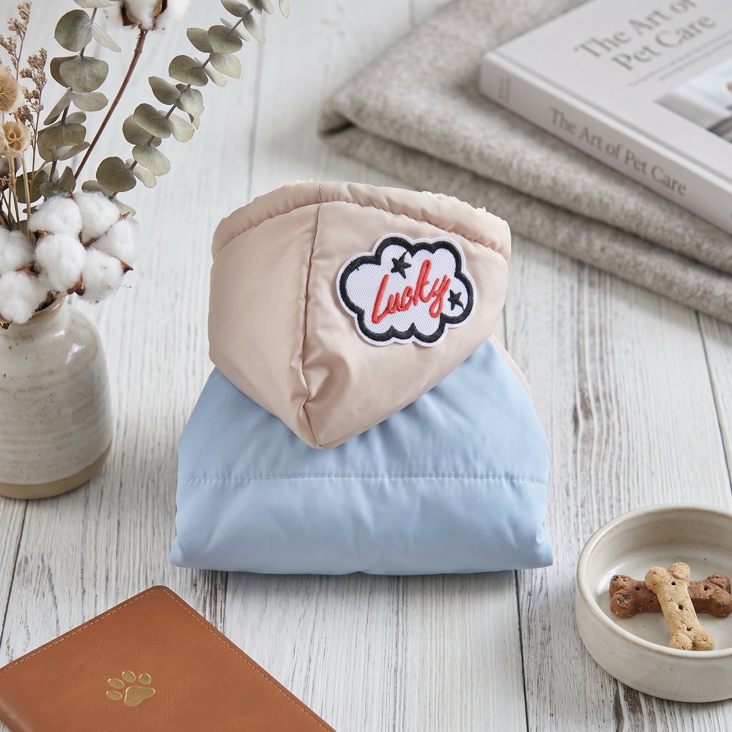 Folded light blue CozyCloud dog jumpsuit with a 'Lucky' cloud patch on the hood, displayed on a rustic wooden table with a dog treat bowl.