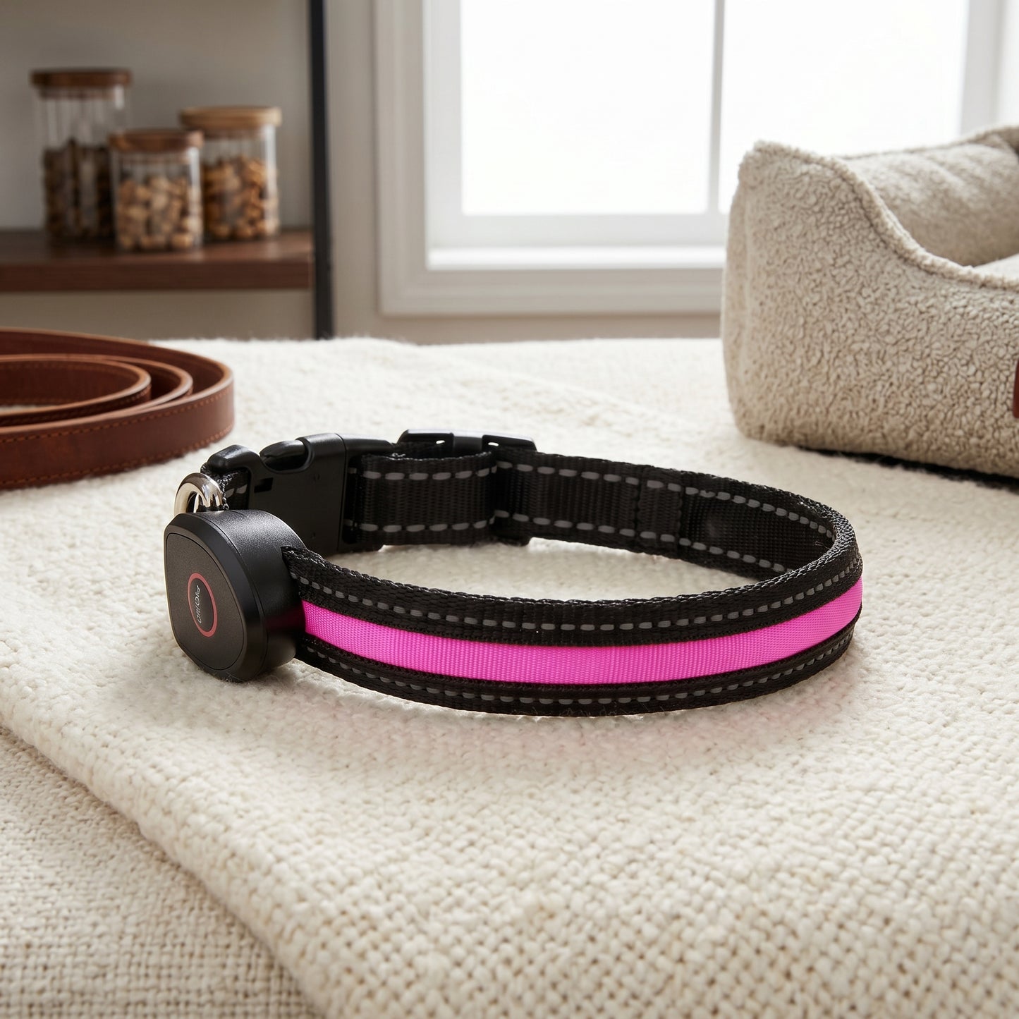 A black dog collar with a bright pink illuminated strip displayed on a white textured blanket next to a rolled leather leash and jars of dog treats.