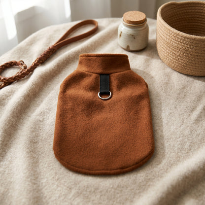 Brown fleece dog sweater displayed on a beige rug next to a woven basket and a leather leash.